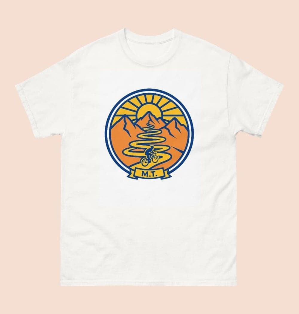 An original crest showing a cyclist riding toward a mountain pass, with a curving trail and rising sun, on a t-shirt (example made with AI)