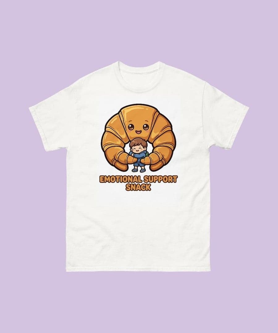 A white T-shirt with a cartoon croissant hugging a little boy and the words Emotional Support Snack on the chest (example)